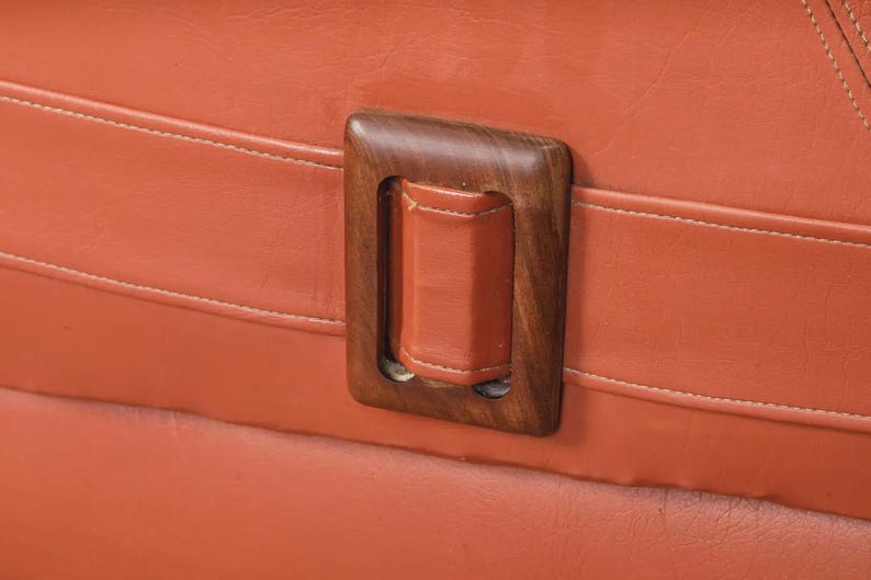 Pu&ograve; includere: Primo piano di una maniglia rettangolare in legno con impugnatura in pelle. La maniglia &egrave; posta su una superficie arancione strutturata con cuciture bianche. La cornice in legno &egrave; marrone scuro.