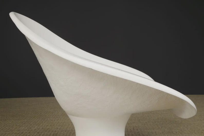 May include: A modern, white, sculptural chair with a curved, petal-like seat and a pedestal base. The chair has a textured surface and a smooth, rounded edge. The design is minimalist and elegant, suitable for contemporary interiors.