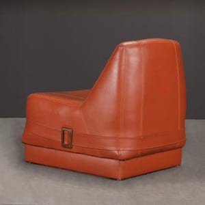 Pu&ograve; includere: Una poltrona in pelle arancione con base rettangolare e schienale alto. La sedia ha un design cucito e un dettaglio con fibbia in metallo sul davanti. La sedia &egrave; ambientata su uno sfondo grigio scuro.