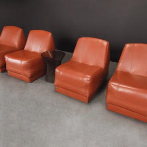 Pu&ograve; includere: Quattro poltrone in pelle arancione-rossa e un tavolino abbinato. Le poltrone hanno un design a basso profilo con una base rettangolare e uno schienale leggermente reclinato. Il tavolino &egrave; scuro e traslucido.
