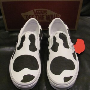 May include: White slip-on trainers with a black cow print pattern. The shoes have white soles and trim. A red tag is attached to one shoe. The Vans box is in the background.