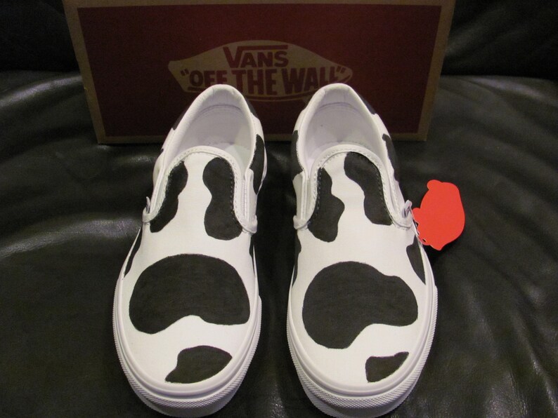May include: White slip-on trainers with a black cow print pattern. The shoes have white soles and trim. A red tag is attached to one shoe. The Vans box is in the background.
