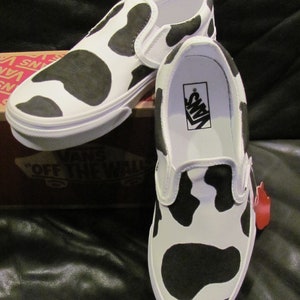 May include: White slip-on trainers featuring a black cow print pattern. The shoes have a white sole and a Vans logo inside. The trainers are displayed on a brown box with the Vans logo.