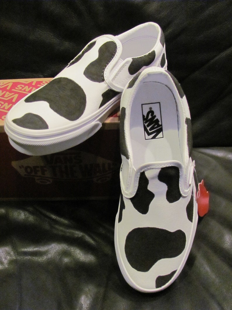 May include: White slip-on trainers featuring a black cow print pattern. The shoes have a white sole and a Vans logo inside. The trainers are displayed on a brown box with the Vans logo.
