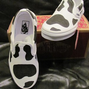 May include: White slip-on trainers featuring a black cow print design. The trainers have a white sole and a black interior. The Vans logo is visible inside one shoe. The trainers are displayed on a brown box.