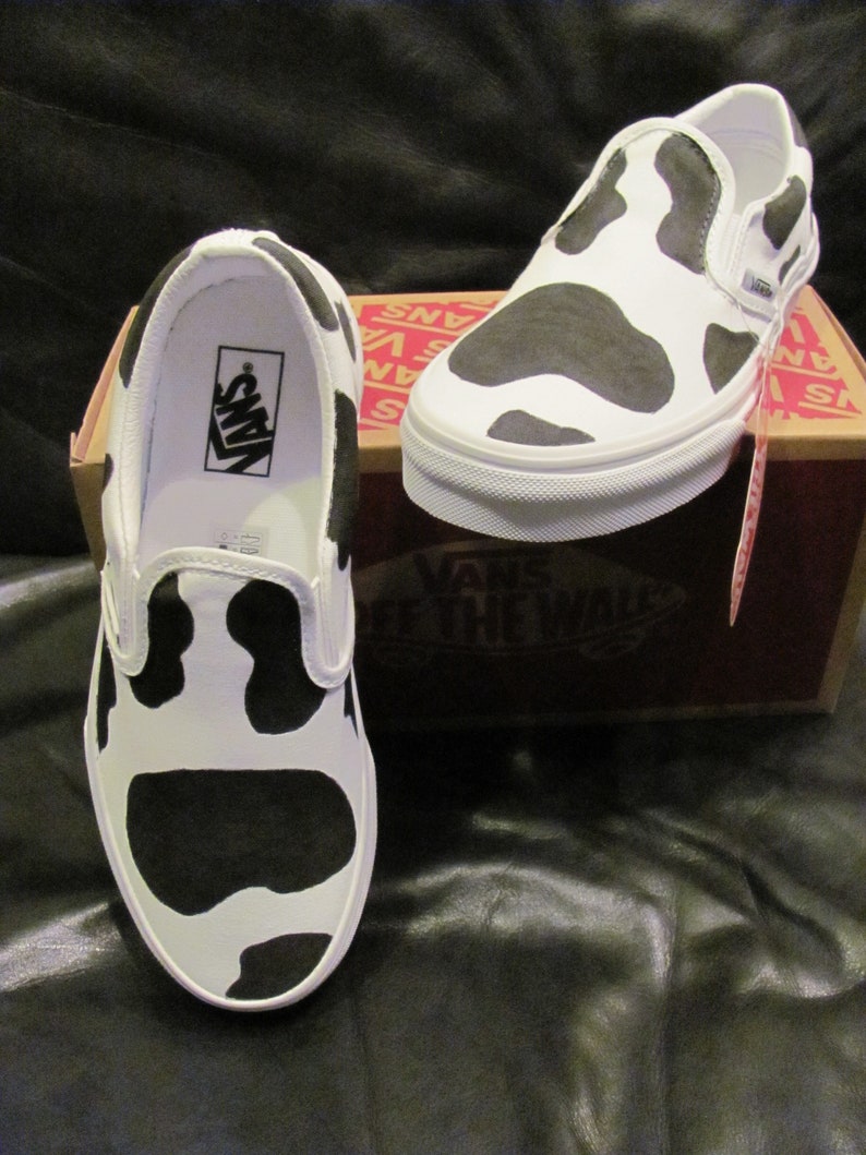 May include: White slip-on trainers featuring a black cow print design. The trainers have a white sole and a black interior. The Vans logo is visible inside one shoe. The trainers are displayed on a brown box.