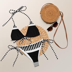 Può includere: Un set bikini a righe bianche e nere con top all'americana e slip con lacci laterali. Il bikini è esposto su un tappetino intrecciato, accanto a una borsa rotonda di paglia e occhiali da sole. Il top e lo slip del bikini presentano un audace motivo a righe.