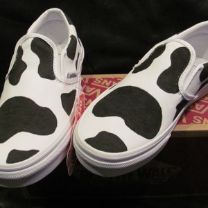 May include: A pair of white slip-on trainers featuring a black cow print design. The shoes have white soles and a Vans logo. They are presented on a Vans shoe box.