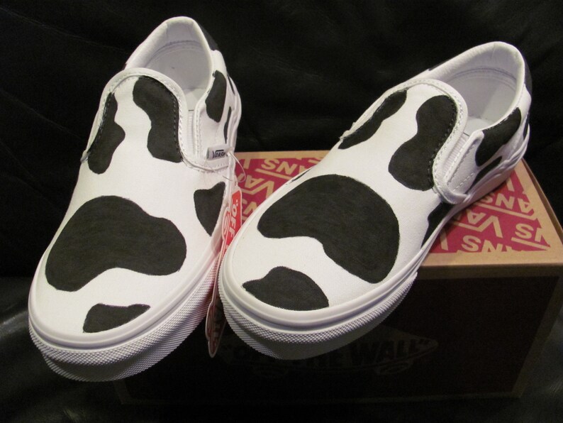 May include: A pair of white slip-on trainers featuring a black cow print design. The shoes have white soles and a Vans logo. They are presented on a Vans shoe box.