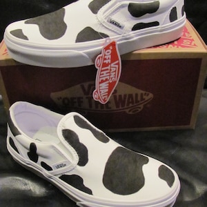 May include: White slip-on trainers featuring a black cow print design. The footwear rests on a brown box with the Vans logo. The trainers have a white sole and the Vans logo on the side. A red tag hangs from the shoe.