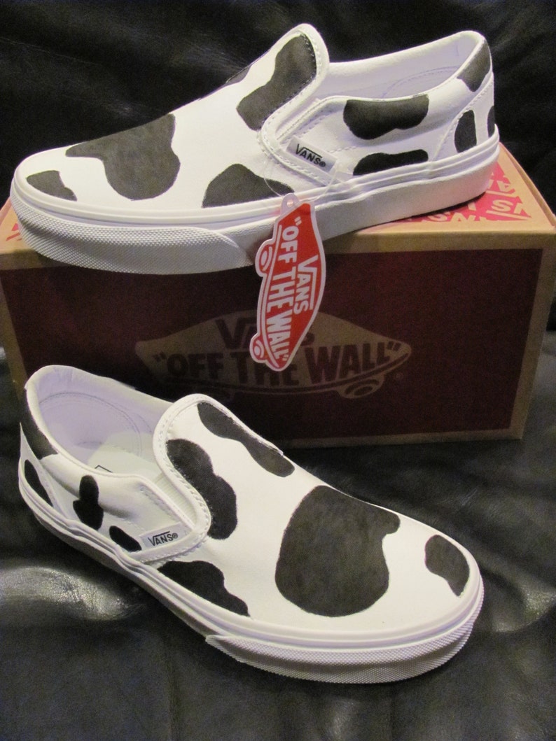 May include: White slip-on trainers featuring a black cow print design. The footwear rests on a brown box with the Vans logo. The trainers have a white sole and the Vans logo on the side. A red tag hangs from the shoe.