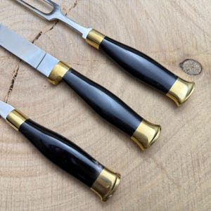 May include: Close-up of a carving set with a knife, fork, and another utensil. The utensils have black handles with gold-colored accents and stainless steel blades. The set is on a wooden surface.