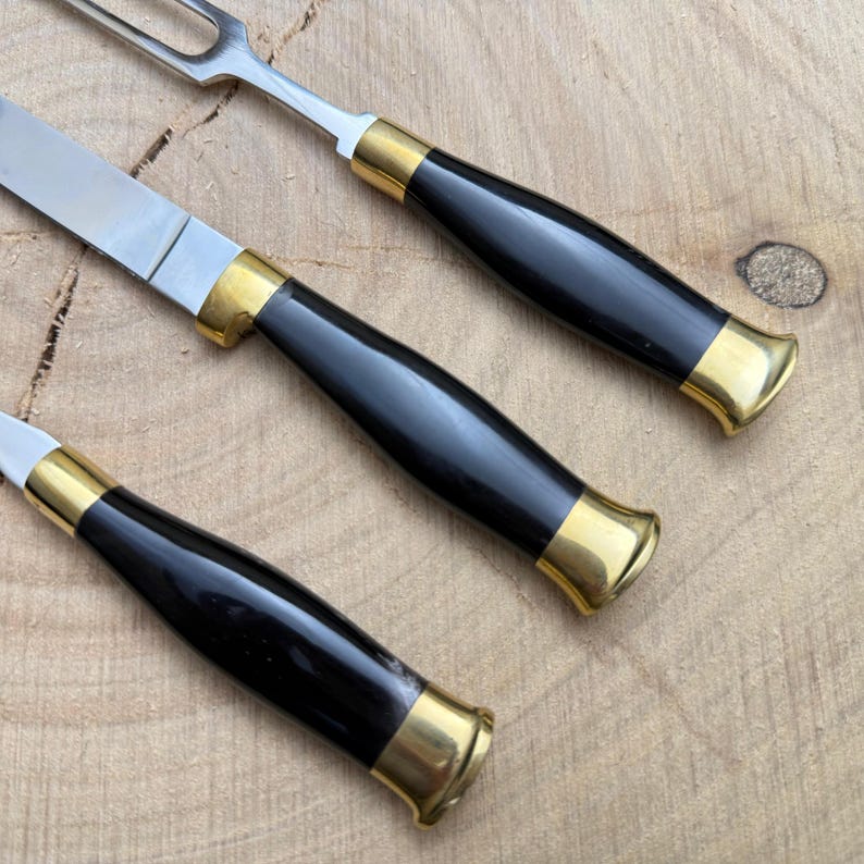 May include: Close-up of a carving set with a knife, fork, and another utensil. The utensils have black handles with gold-colored accents and stainless steel blades. The set is on a wooden surface.