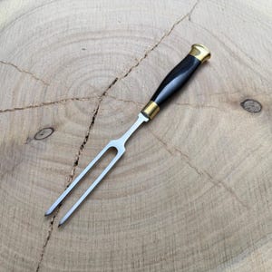 May include: A stainless steel carving fork with a black handle and brass accents. The two-pronged fork is designed for handling and serving roasted meats. The handle is a glossy black with a brass bolster and end cap.