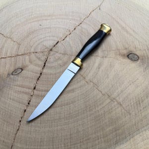 May include: A handcrafted knife with a polished steel blade and a black handle. The handle is accented with brass-colored metal at both ends. The knife is displayed on a light brown wooden surface, showcasing its sharp design.