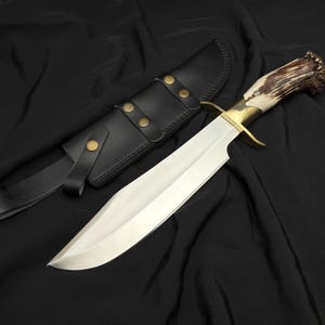 K&ouml;nnte beinhalten: Ein gro&szlig;es, poliertes Stahlmesser mit einem Messing-Handschutz und einem Griff aus Knochen und Geweih. Das Messer wird von einer schwarzen Lederscheide mit Messingnieten und einer G&uuml;rtelschlaufe begleitet. Die Klinge ist lang und scharf.