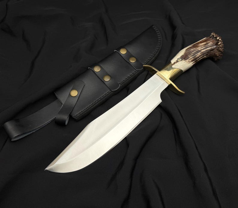 K&ouml;nnte beinhalten: Ein gro&szlig;es, poliertes Stahlmesser mit einem Messing-Handschutz und einem Griff aus Knochen und Geweih. Das Messer wird von einer schwarzen Lederscheide mit Messingnieten und einer G&uuml;rtelschlaufe begleitet. Die Klinge ist lang und scharf.