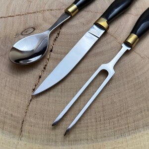 May include: Close-up of a stainless steel spoon, knife, and fork set. The utensils have black handles with gold-colored accents. The knife has a long, sharp blade. The set is displayed on a wooden surface.
