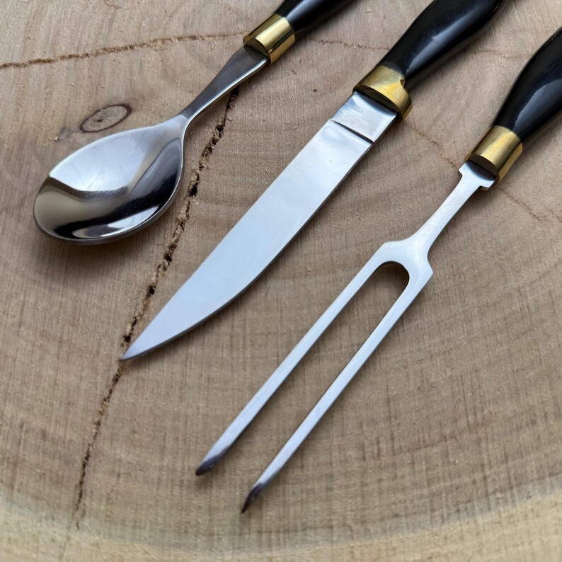 May include: Close-up of a stainless steel spoon, knife, and fork set. The utensils have black handles with gold-colored accents. The knife has a long, sharp blade. The set is displayed on a wooden surface.