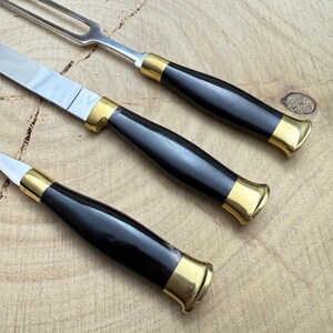 May include: A close-up of a carving set with a knife, carving fork, and a smaller knife. The handles are black with gold-colored metal accents. The blades are silver and the set is on a wooden surface.
