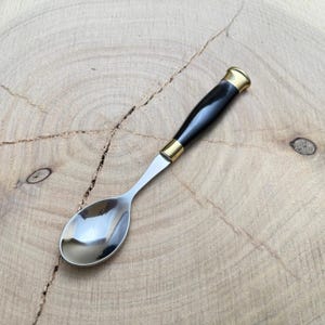 May include: A small spoon with a polished stainless steel bowl and a black handle. The handle is accented with gold-colored metal bands at the base and the end. The spoon is designed for serving or eating.