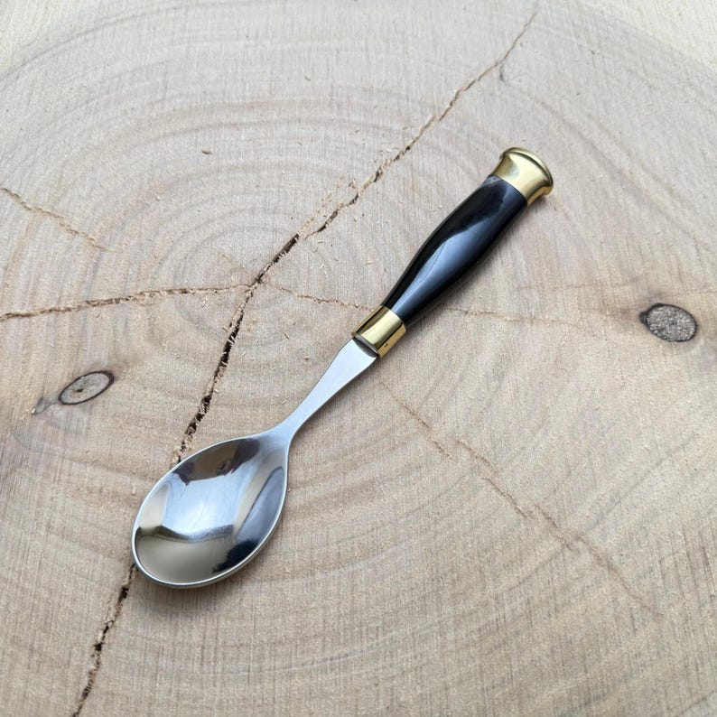 May include: A small spoon with a polished stainless steel bowl and a black handle. The handle is accented with gold-colored metal bands at the base and the end. The spoon is designed for serving or eating.