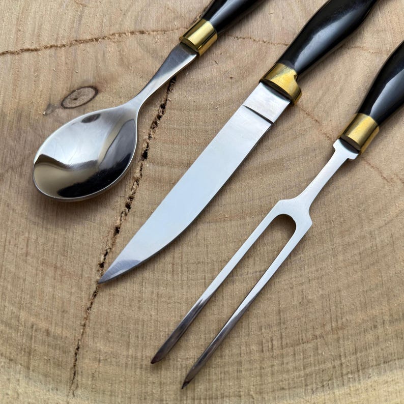 May include: Close-up of a set of three stainless steel serving utensils with black handles and gold-colored accents. The set includes a spoon, a knife, and a two-pronged fork. The utensils are arranged on a wooden surface.