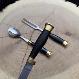 May include: A set of three stainless steel utensils: a knife, a fork, and a spoon. The knife and fork have black handles with gold-colored accents. The spoon has a white handle with gold-colored accents. The utensils are arranged on a wooden surface.