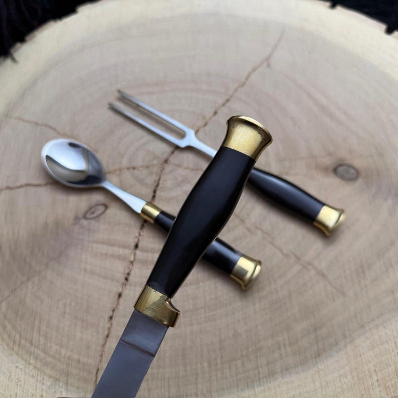 May include: A set of three stainless steel utensils: a knife, a fork, and a spoon. The knife and fork have black handles with gold-colored accents. The spoon has a white handle with gold-colored accents. The utensils are arranged on a wooden surface.