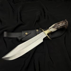 K&ouml;nnte beinhalten: Ein gro&szlig;es, poliertes Stahlmesser mit einem Knochen- und Geweihgriff, einem Messingb&uuml;gel und einer schwarzen Lederscheide. Die Klinge ist breit und scharf, und der Griff ist f&uuml;r den Halt strukturiert. Das Messer ist f&uuml;r den Au&szlig;enbereich konzipiert.