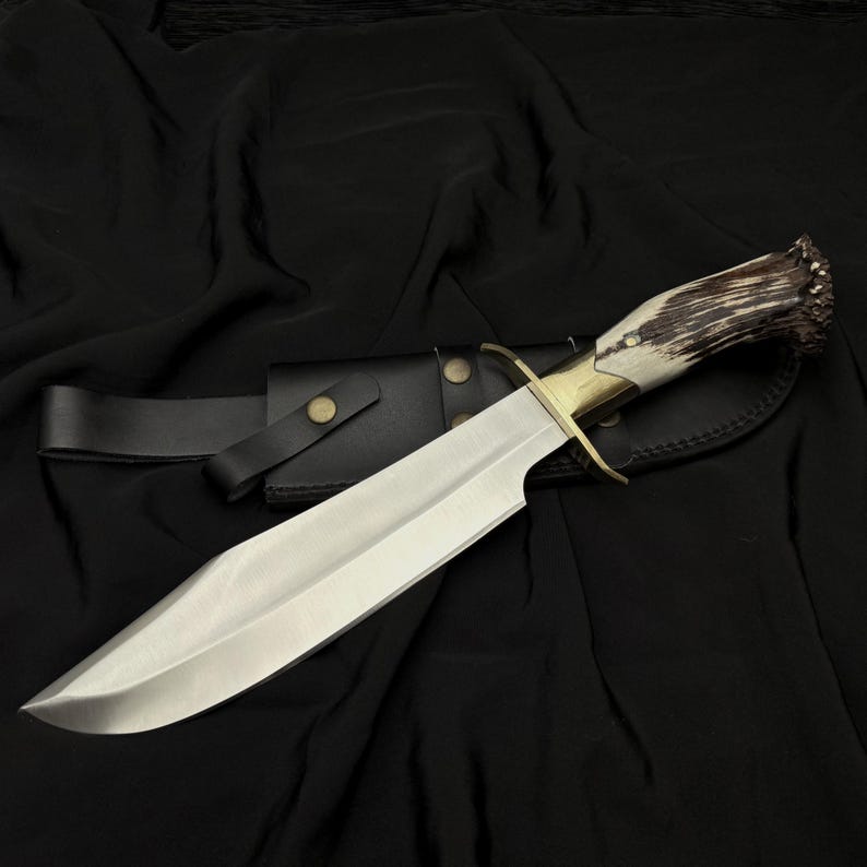 K&ouml;nnte beinhalten: Ein gro&szlig;es, poliertes Stahlmesser mit einem Knochen- und Geweihgriff, einem Messingb&uuml;gel und einer schwarzen Lederscheide. Die Klinge ist breit und scharf, und der Griff ist f&uuml;r den Halt strukturiert. Das Messer ist f&uuml;r den Au&szlig;enbereich konzipiert.