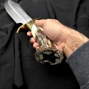K&ouml;nnte beinhalten: Ein gro&szlig;es Jagdmesser mit polierter Stahlklinge und Messingb&uuml;gel. Der Griff besteht aus Geweihmaterial mit sternf&ouml;rmigem Ende. Das Messer wird in einer Hand vor dunklem Hintergrund gehalten.