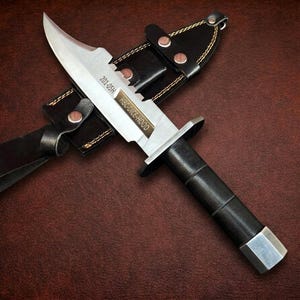 May include: A fixed-blade knife with a silver-toned steel blade featuring the text 201-05H and HBC-DICE-HOOD, a black cylindrical handle, and a silver pommel. The knife is accompanied by a black leather sheath with copper rivets and yellow stitching.