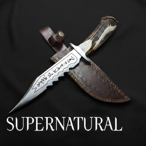 May include: A silver knife with a serrated edge and etched text, resting on a brown leather sheath. The handle is made of antler. The word "SUPERNATURAL" is displayed in white text below the knife.