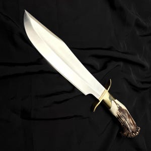 K&ouml;nnte beinhalten: Ein gro&szlig;es, poliertes Stahlmesser mit gebogener Klinge. Das Messer hat einen Messingknauf und einen Griff aus Knochen- und Geweihmaterial. Die Klinge ist etwa 38 cm lang. Das Messer wird vor einem schwarzen Hintergrund pr&auml;sentiert.