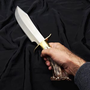 K&ouml;nnte beinhalten: Ein gro&szlig;es, poliertes Stahlmesser mit breiter Klinge und Messing-Handschutz. Der Griff besteht aus braunem und beigem Geweihmaterial. Das Messer wird in einer Hand vor schwarzem Hintergrund gehalten.