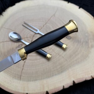 May include: A knife, fork, and spoon set with black handles and gold-colored accents. The knife blade is silver. The utensils are arranged on a light brown wooden surface.