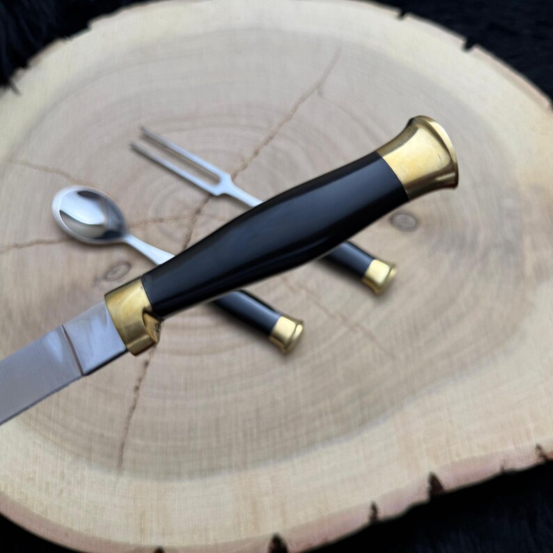 May include: A knife, fork, and spoon set with black handles and gold-colored accents. The knife blade is silver. The utensils are arranged on a light brown wooden surface.