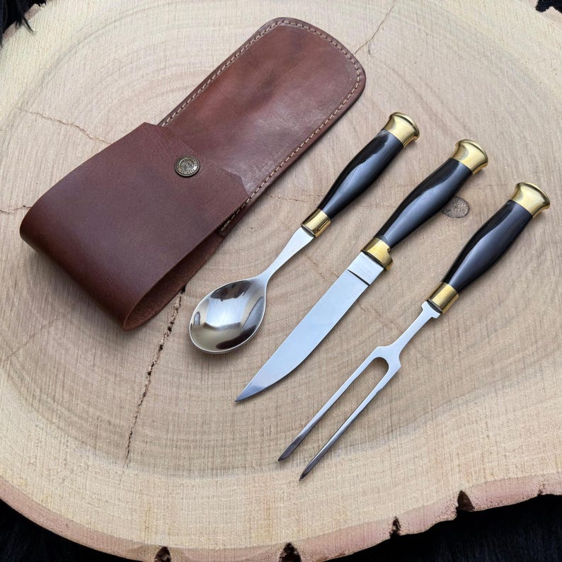 May include: A three-piece cutlery set with a brown leather carrying case. The set includes a spoon, knife, and fork, each with black handles, gold accents, and stainless steel blades. The case has a snap closure.