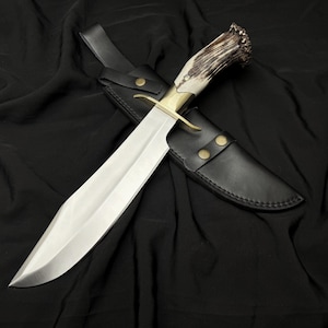 May include: A large, polished steel knife with a brass guard and a handle made of antler. The knife is resting in a black leather sheath with brass rivets. The blade is long and sharp, and the overall design suggests a hunting or survival knife.