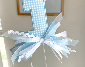 Puede incluir: Un adorno para pastel con el número uno en cuadros vichy azul claro y blanco. La base del número está decorada con un conjunto de cintas en azul claro, blanco y texturas translúcidas, incluyendo un borde ondulado, sujeto a un palo de plástico.