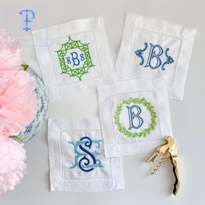 May include: Four white linen napkins with embroidered initials. The napkins feature different embroidery designs, including a monogram, a wreath, and a geometric pattern. The initials are EBS, B, and S.