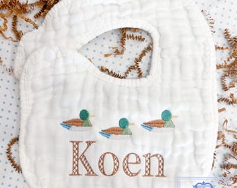 Op de afbeelding: Witte katoenen baby-slabbetje met een geborduurde naam "Koen" in bruin en drie groene eendjes.