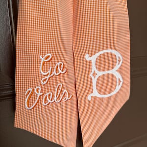 May include: Orange and white checkered fabric with the embroidered text "Go Vols" in orange script and a white letter "B". The fabric is folded and hanging.