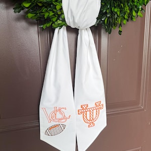 May include: A green wreath with a white fabric bow and two white fabric streamers. The streamers have orange and white embroidered designs, including a football and collegiate logos. The wreath is hanging on a brown door.