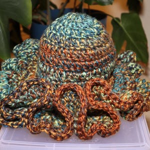 May include: A handmade crochet hat with a matching octopus-like design. The hat and tentacles are made with yarn in shades of teal, orange, and brown. The hat is sitting on a clear plastic container.