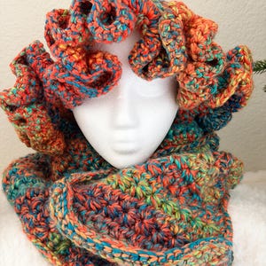 May include: A colorful, handmade crochet cowl and head wrap set. The set features a vibrant mix of orange, blue, and green yarn. The cowl is thick and textured, while the head wrap has a unique, ruffled design. This cozy set is perfect for cold weather.