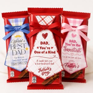 May include: Three red chocolate bars decorated with tie-shaped paper labels. The left label is blue plaid with "World's BEST DAD". The centre is red plaid with "DAD, You're One of a Kind". The right is pink plaid with "DAD, You're the Sweetest".