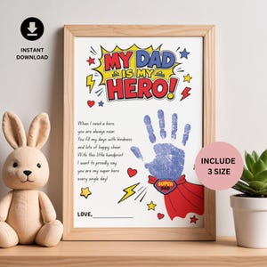 Könnte beinhalten: Ein gerahmter Druck mit dem Text MY DAD IS MY HERO im Comic-Stil. Er enthält eine blaue Handabdruck-Grafik mit einem roten Superheldenumhang und dem Text SUPER DAD auf dem Emblem. Links steht ein Gedicht mit einer leeren Zeile für eine Unterschrift.