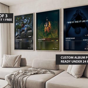 May include: Three framed album art posters with black borders are displayed on a wall. Each poster features a different album cover design with the album name and artist information. The posters are above a light-coloured sofa.